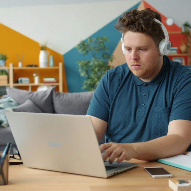 Student in headphones working on a laptop at a desk with an open notebook