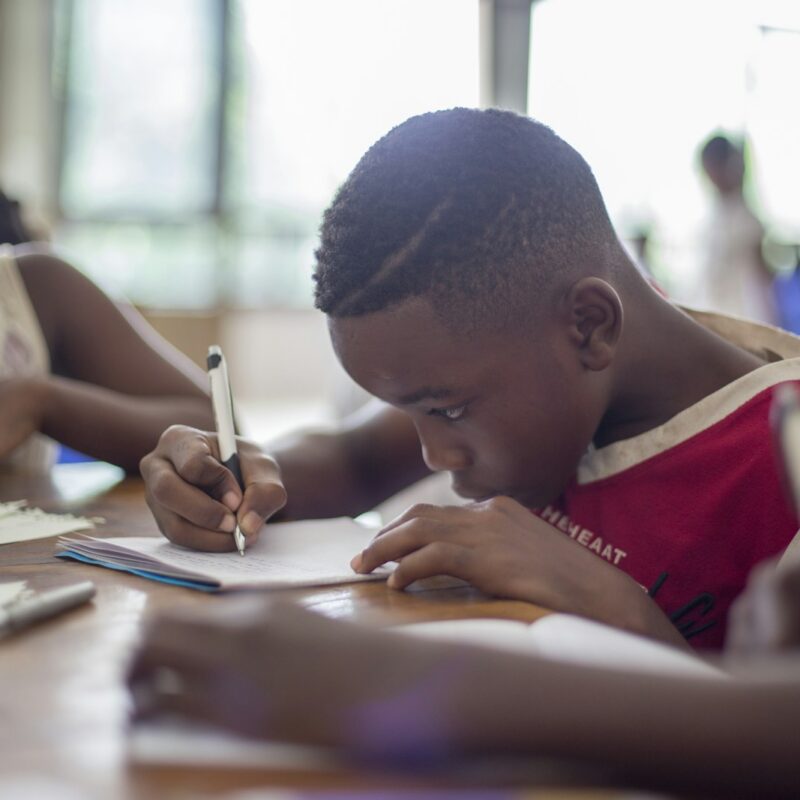A student bent over a sheet of paper, writing with a pen at a wooden table, with a classmate reading beside him