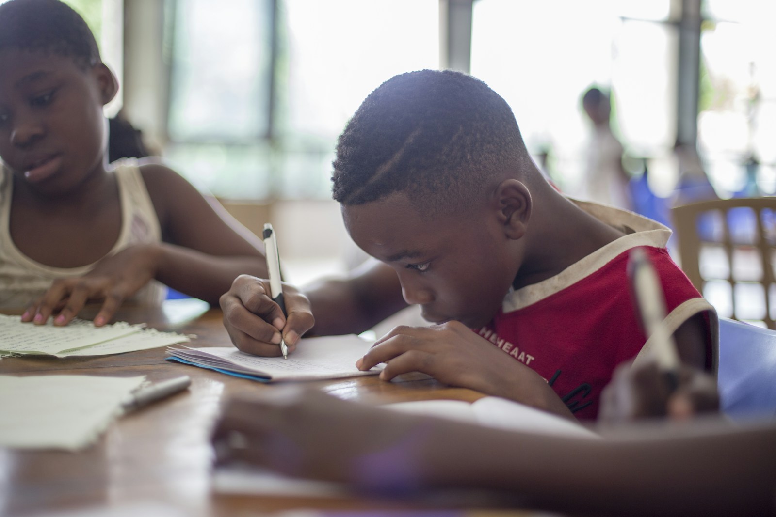A student bent over a sheet of paper, writing with a pen at a wooden table, with a classmate reading beside him